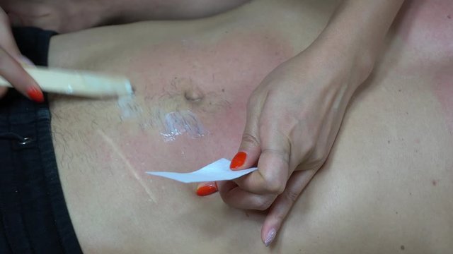 close up of a spatula spreading a thin layer of hot wax on the man's abdomen and navel. A strip is placed over the waxed area, pressed down and pulled firmly removing the unwanted hair 