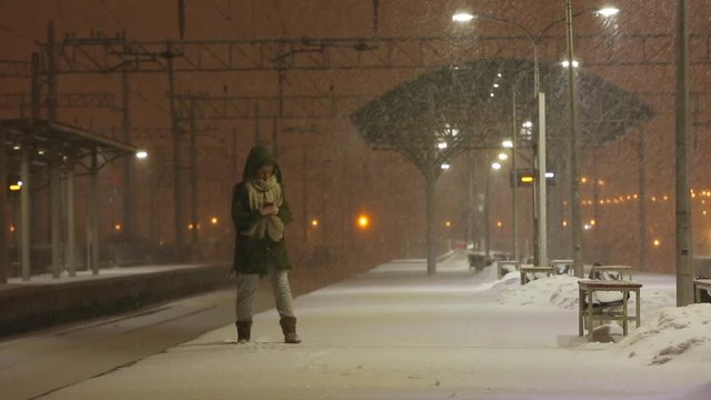 Young woman in hood on empty railway platform in blizzard waiting for a train. Female is waiting for the train in bad weather in the evening, checking smartphone, strong wind.