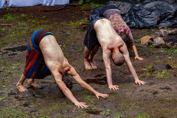Diverse people enjoy spiritual gathering Two shirtless men are seen practicing yoga in a muddy forest clearing outdoors, seen in downward-facing dog pose (adho mukha svanasana). With copy-space.