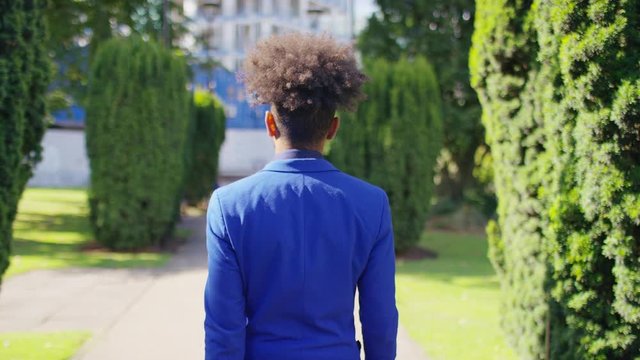 Camera follows behind a hip male in a suit as he walks through a city garden, in slow motion