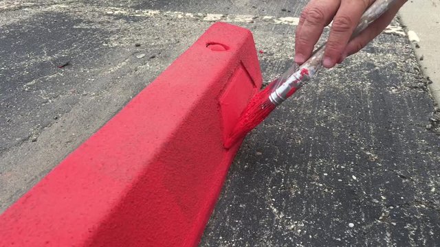 Occupational painter applying red paint onto parking stop in lot before dipping brush into bucket up close.