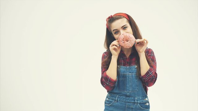 Lovely pinup girl is hiding her mouth behind big pink doughnut, making big eyes, smiling, raising eyebrows.