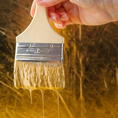 Hand holds a paintbrush with dripping golden paint over background in gold colors.