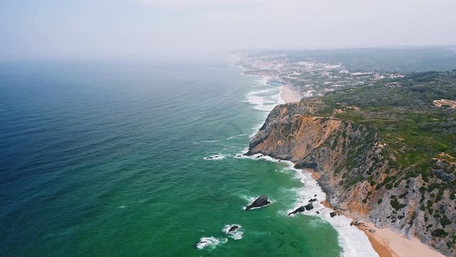 4K Aerial view of Sintra coastline, Portugal. Praia da Adraga beach and atlantic coast with white ocean waves rolling towards the shore
