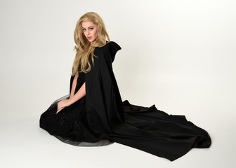 full length portrait of blonde girl wearing long black flowing cloak, sitting on the floor  with  a...
