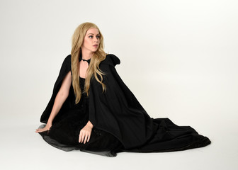 full length portrait of blonde girl wearing long black flowing cloak, sitting on the floor  with  a...
