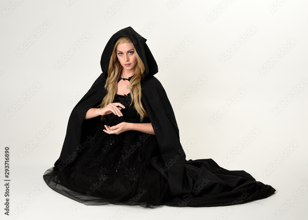 Sticker full length portrait of blonde girl wearing long black flowing cloak, sitting on the floor  with  a white studio background.