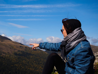 The girl shows a finger to the distance towards the mountains. A girl in a denim jacket, sunglasses and a scarf on her head