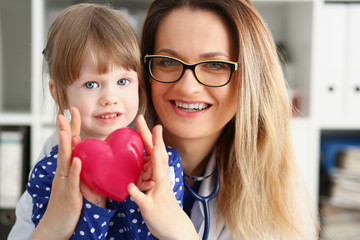 Female doctor and happy little child hold in arms red toy heart closeup. Cardio therapeutist...