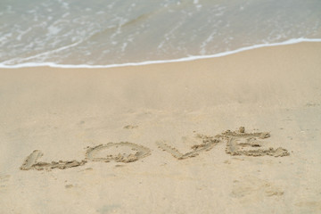 Love written on sand, in the background soft waves with foam of ocean on the sandy beach.