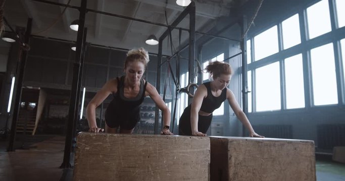 Slow motion two young beautiful female athletes working out, doing push-ups and clapping together in atmospheric gym.