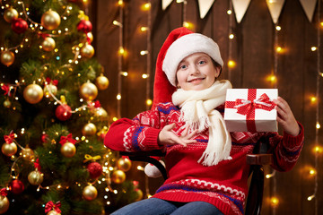 Cheerful santa helper girl with gift box sitting indoor near decorated xmas tree with lights, dressed in red sweater - Merry Christmas and Happy Holidays!