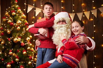 Santa Claus and child boy and girl posing together indoor near decorated xmas tree with lights, they talking, smiling and accepting gifts - Merry Christmas and Happy Holidays!