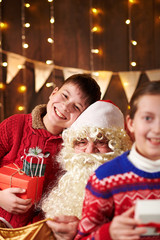 Santa Claus opens a bag and gives gifts to children, sitting indoor near decorated xmas tree with lights - Merry Christmas and Happy Holidays!