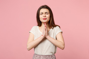 Young woman in casual light clothes posing isolated on pastel pink wall background, studio portrait. People lifestyle concept. Mock up copy space. Holding hands folded in prayer, keeping eyes closed.