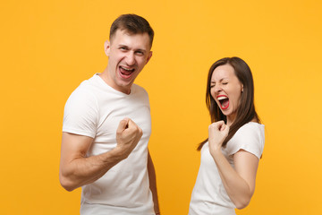 Overjoyed young couple friends guy girl in white empty blank t-shirts posing isolated on yellow orange background. People lifestyle concept. Mock up copy space. Clenching fists like winners screaming.