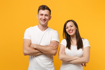 Cheerful young couple two friends guy girl in white empty blank design t-shirts posing isolated on yellow orange wall background. People lifestyle concept. Mock up copy space. Holding hands crossed.