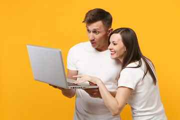 Smiling young couple two friends guy girl in white blank t-shirts posing isolated on yellow orange background. People lifestyle concept. Mock up copy space. Holding, working on laptop pc computer.