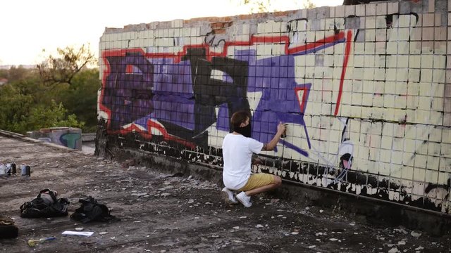 Backside view of a young caucasian guy in casual clothes creatively works with a outdoors bricked wall, applying spray on a surface on the top roof. In process of creation. Urban, youth, creativety