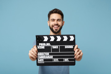 Young smiling man in casual clothes posing isolated on blue wall background, studio portrait. People lifestyle concept. Mock up copy space. Holding in hands classic black film making clapperboard.
