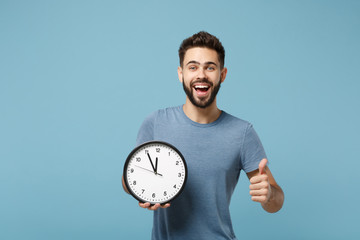 Young excited funny man in casual clothes posing isolated on blue background, studio portrait. People sincere emotions lifestyle concept. Mock up copy space. Holding in hands clock, showing thumb up.