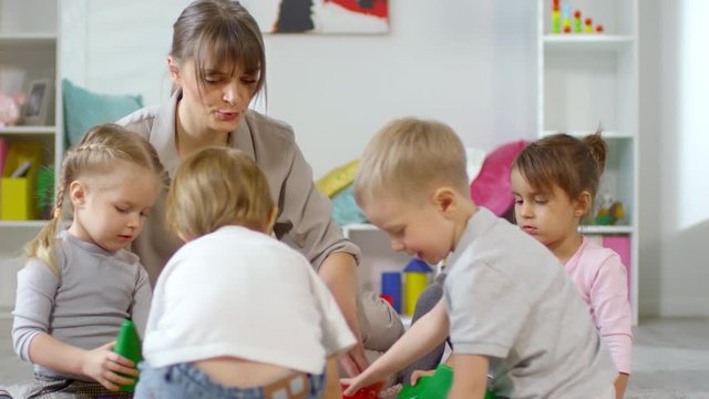 Female teacher sitting on the floor with kids and explaining them how to play educational game with colorful toy blocks in daycare playroom