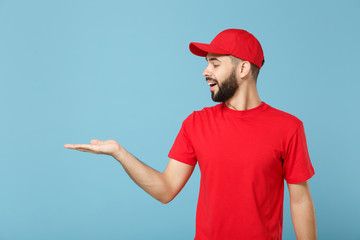 Delivery man in red uniform workwear isolated on blue wall background, studio portrait. Professional male employee in cap t-shirt print working as courier dealer. Service concept. Mock up copy space.