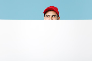 Delivery man in red workwear hold big white empty billboard isolated on blue background, studio portrait. Male employee in cap t-shirt working as courier dealer. Service concept. Mock up copy space.