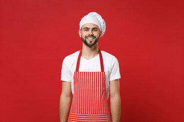 Smiling young bearded male chef cook or baker man in striped apron white t-shirt toque chefs hat posing isolated on bright red background, studio portrait. Cooking food concept. Mock up copy space.