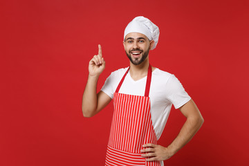 Bearded male chef cook baker man in striped apron white t-shirt toque chefs hat posing isolated on red background. Cooking food concept. Mock up copy space. Hold index finger up with great new idea.