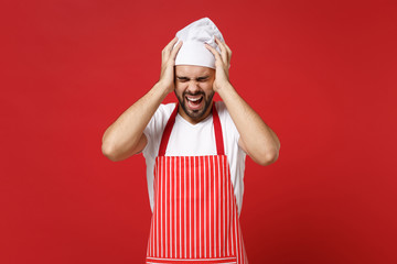 Bearded male chef cook or baker man in striped apron toque chefs hat posing isolated on red background. Cooking food concept. Mock up copy space. Screaming, keeping eyes closed, putting hands on head.