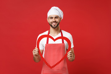 Smiling young bearded male chef cook or baker man in striped apron white t-shirt toque chefs hat isolated on red wall background. Cooking food concept. Mock up copy space. Holding big wooden heart.