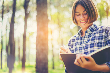 Pretty girl with notebook and pen make a short note while travel education along path of a big forest.