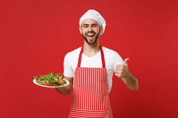 Joyful young male chef cook or baker man in striped apron toque chefs hat posing isolated on red background. Cooking food concept. Mock up copy space. Hold plate with vegetable salad showing thumb up.