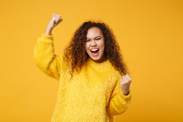 Overjoyed young african american girl in fur sweater posing isolated on yellow orange background in studio. People sincere emotions lifestyle concept. Mock up copy space. Clenching fists like winner.