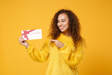 Smiling young african american girl in fur sweater posing isolated on yellow orange background studio portrait. People lifestyle concept. Mock up copy space. Pointing index finger on gift certificate.