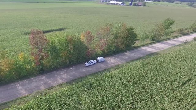 Aerial View of a white Dodge Avenger car hauling a small retro teardrop camping trailer down a dirt road in the country.