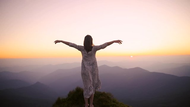 Woman hiker open arms at mountain peak at sunset. The camera moves around girl in a long dress fluttering in the wind at the top of a mountain