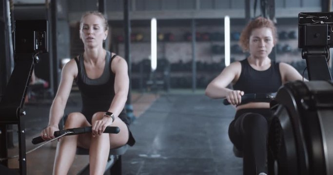 Active and healthy lifestyle, young athletic happy Caucasian women work out together on rowing gym machines slow motion.