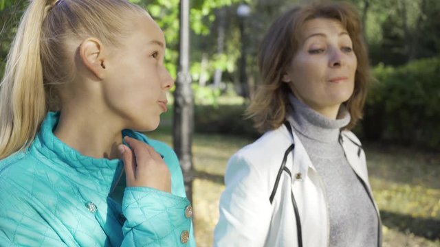 Close-up of a charming caucasian brunette woman talking to the teenage blond girl. Grandma spending time with her granddaughter in the sunny autumn park. Good relashionship between generations.