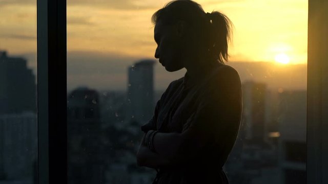 Sad, desperate woman by window at home during sunset