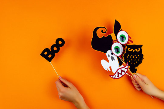 Female hands are holding paper photo props on orange background. Scary ghost, owl, vampire smile, teeth, blood eyes, witch hat on canvas. Party carnival, accessories for celebrating happy halloween.