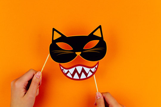 Funny face of monster with cat mask and vampire smile on orange background. Female hands are holding paper photo props on canvas. Party carnival accessories for celebration happy halloween.