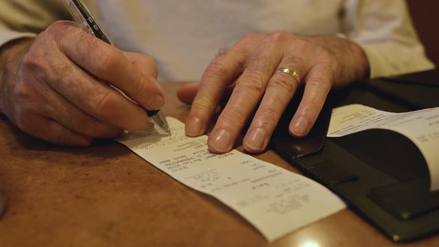 Close Up of a Married Male Senior Citizen's Hands Signing the Bill at a Restaurant, while wearing a gold ring and a white dress shirt