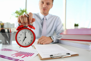 Male hand on the alarm clock a red color stands in the office on the table showing seven o'clock in...