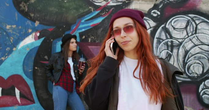 Caucasian young stylish hipster girl in sunglasses and hat talking on the mobile phone while standing outdoor with a graffity wall and another girl on the background. Close up.