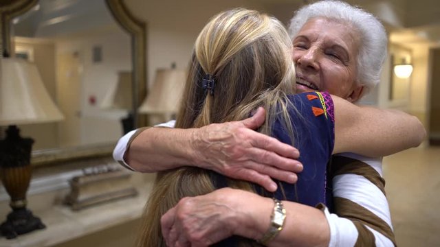 Elderly woman smiles as blonde woman enters and they hug.