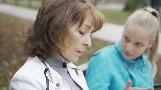 Two caucasian women of different ages sitting in the sunny park and talking. Irritated blond teen complaining about something to the older brunette woman. Focused on the foreground.