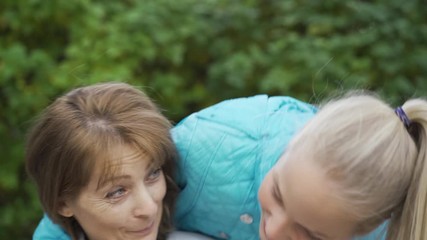 Young blond teen creeping to a brunette senior woman from the back and closing her eyes with hands. Cheerful granddaughter surprising her grandma. Family meeting outdoors.