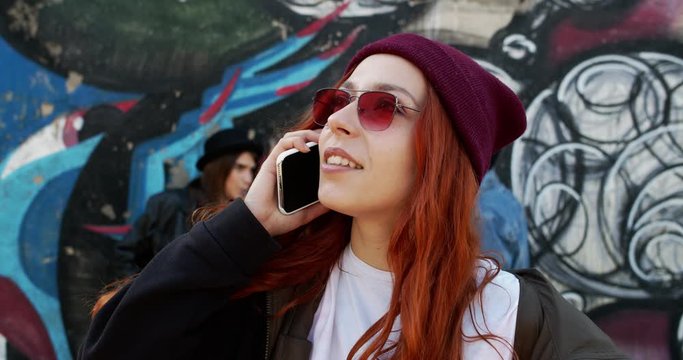 Close up of the young red-haired stylish Caucasian hpster girl in sunglasses and hat speaking on the phone on the graffity wall and couple talking background. Outdoors.